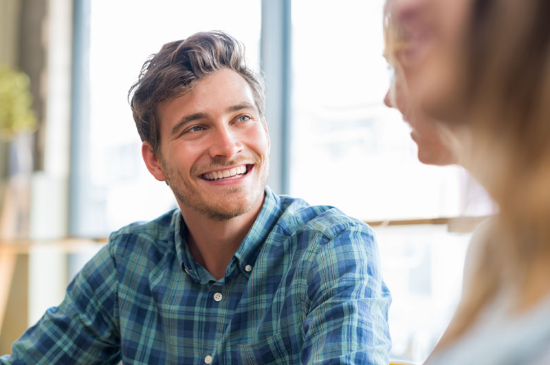 Closeup of cheerful young man in conversation with woman in cafeteria. Happy young couple talking and smiling sitting at table. Handsome guy smiling with his friends.